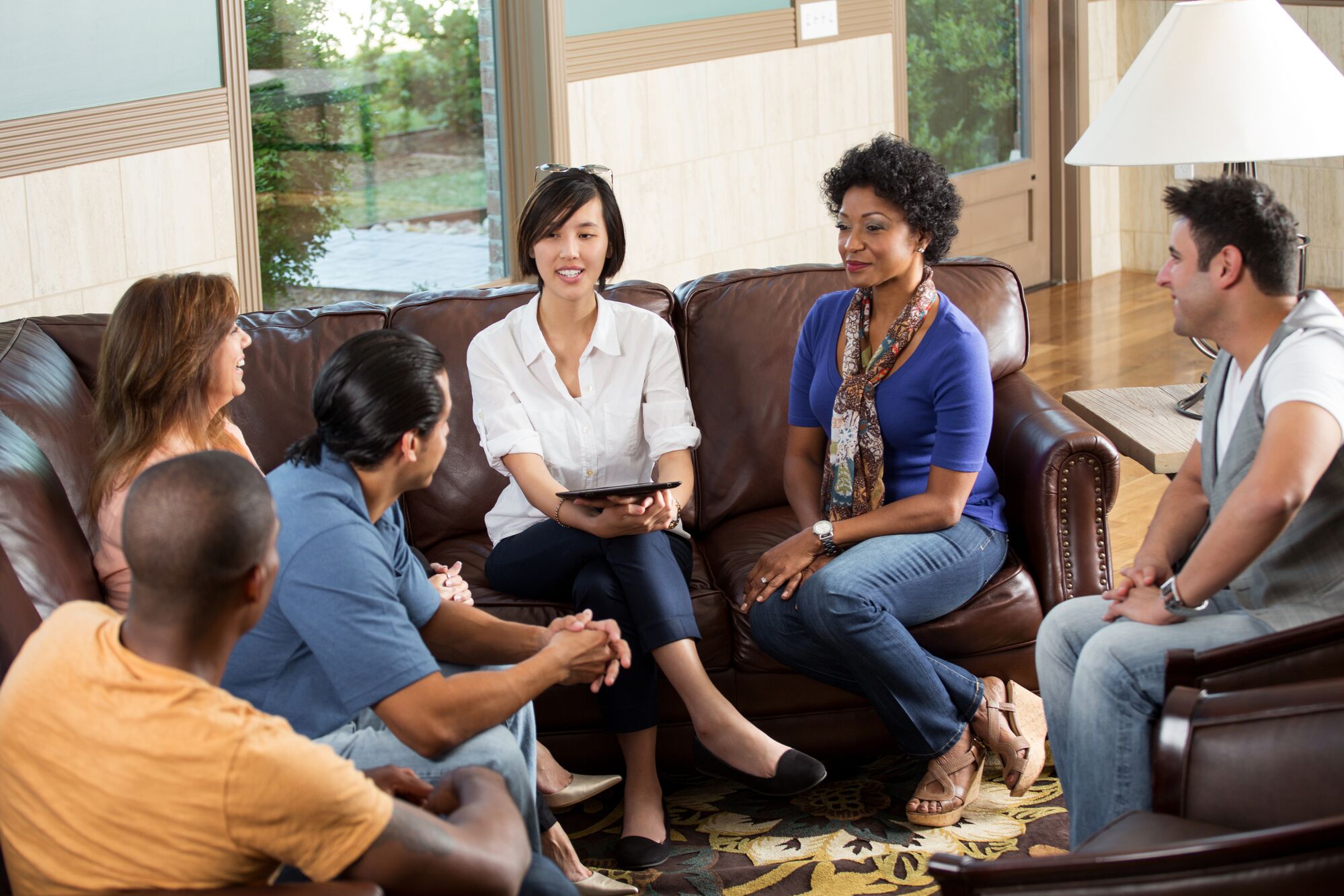 A diverse group of five adults sits in a cozy living room, engaging in conversation. One woman holds a tablet, facilitating the discussion.